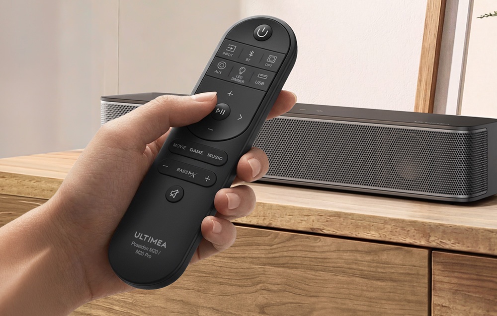 Ένα τηλεχειριστήριο για τη soundbar, κρατημένο στο χέρι, με τη soundbar να βρίσκεται πάνω σε μια ξύλινη συρταριέρα στο βάθος