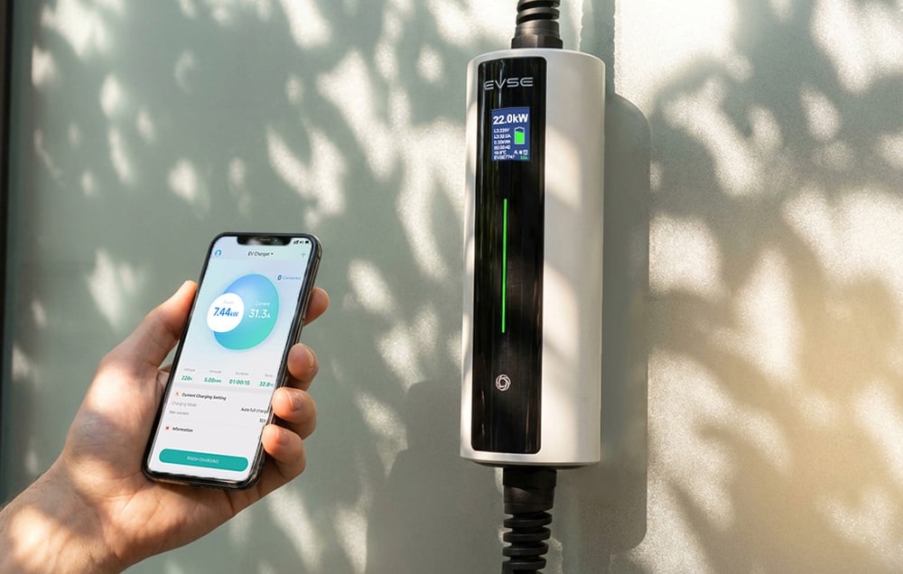 Ένα smartphone στο χέρι με την εφαρμογή ανοιχτή· ο φορτιστής ηλεκτρικών οχημάτων βρίσκεται στα δεξιά