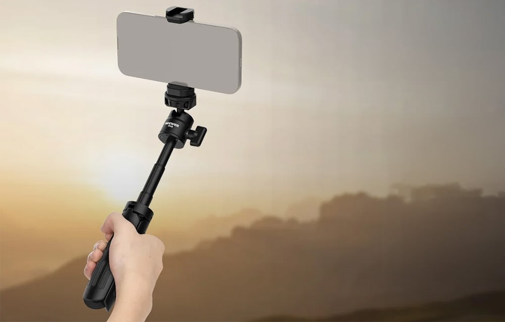 Ένα χέρι που κρατά ένα τηλεσκοπικό selfie stick με βάση για smartphone κατά τη διάρκεια εγγραφής με φόντο το ηλιοβασίλεμα.