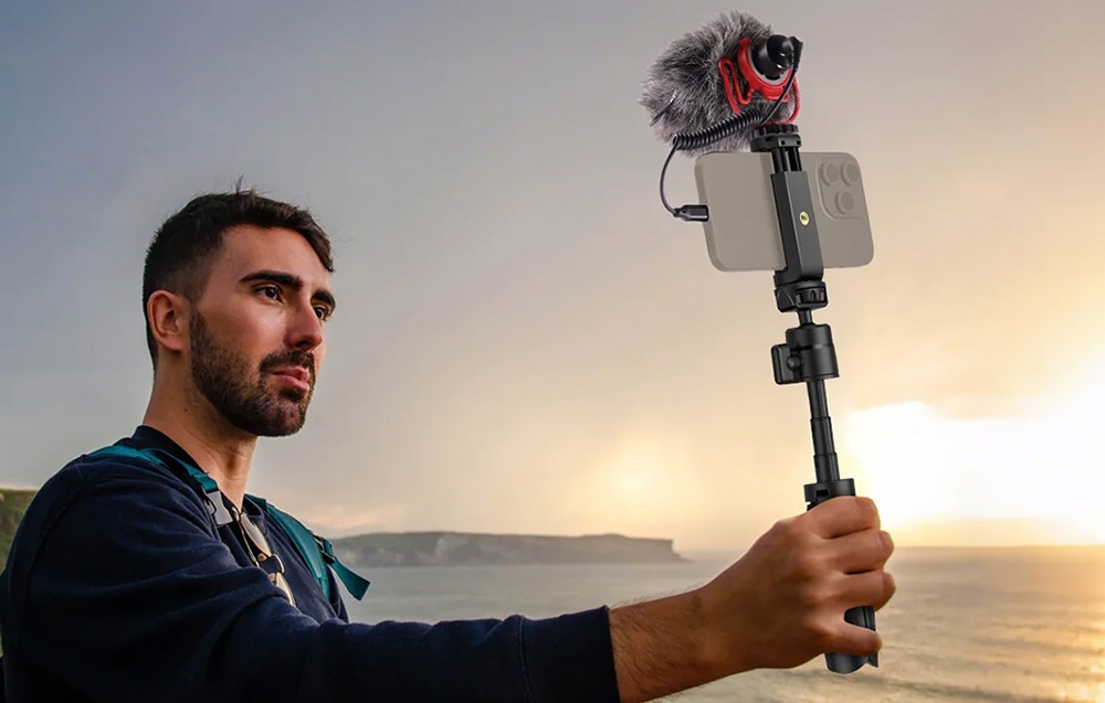 Ένας άνδρας που εγγράφει ένα vlog με ένα smartphone και ένα εξωτερικό μικρόφωνο τοποθετημένα σε ένα τηλεσκοπικό τρίποδο δίπλα στη θάλασσα.