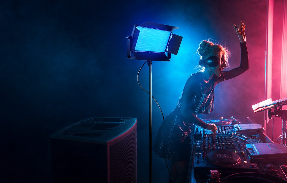 Μια DJ παίζει σε μια κονσόλα μουσικής σε ένα σκοτεινό στούντιο που φωτίζεται από μπλε και ροζ φως από μια λάμπα.