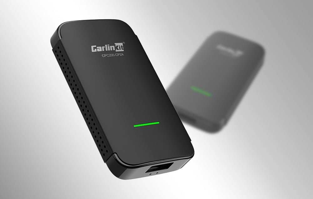 Μαύρος προσαρμογέας CarlinKit CPC200-CP2A με πράσινο LED, όπως φαίνεται σε ανοιχτόχρωμο φόντο, με μια θολή δεύτερη συσκευή στο παρασκήνιο