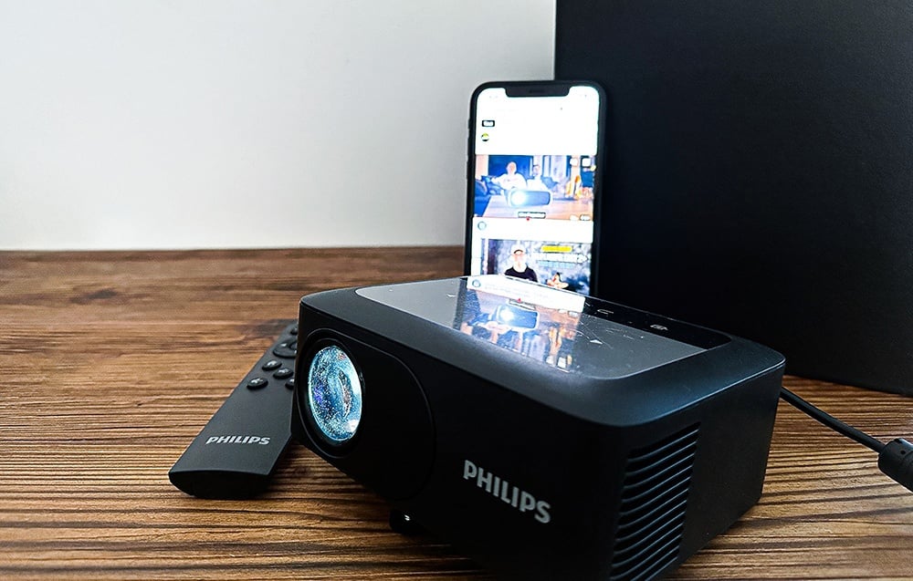 Ένας μαύρος προβολέας PHILIPS πάνω σε ξύλινη επιφάνεια τραπεζιού, με ένα τηλεχειριστήριο PHILIPS και ένα smartphone που προβάλλει μια ταινία δίπλα του