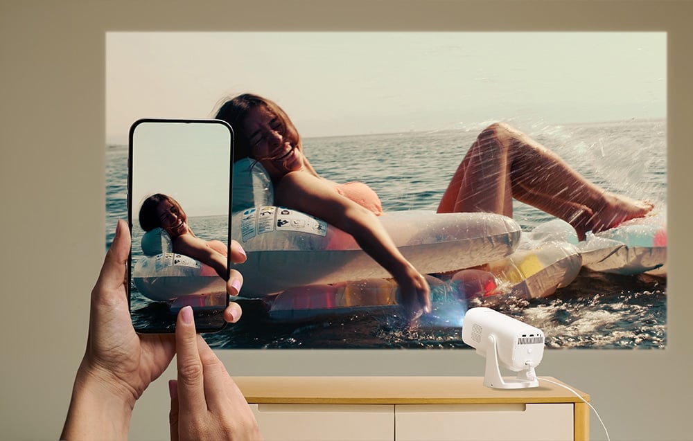 Χέρια κρατούν ένα smartphone που δείχνει μια γυναίκα στο νερό· η ίδια εικόνα προβάλλεται από έναν λευκό προβολέα που βρίσκεται πάνω σε ένα ντουλάπι