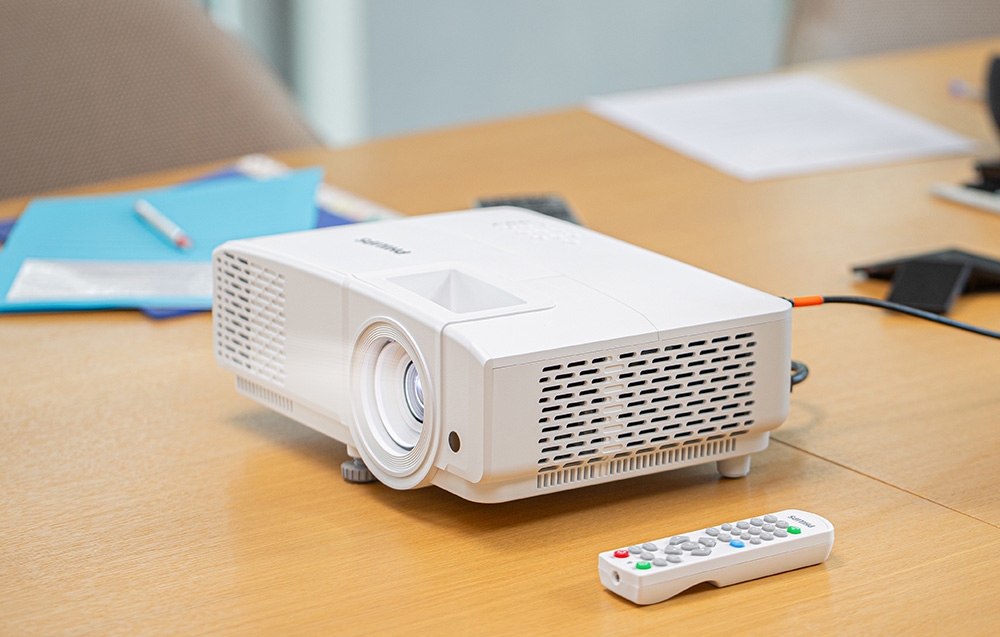 Ένας λευκός προβολέας PHILIPS βρίσκεται πάνω σε ένα ξύλινο τραπέζι συνεδριάσεων δίπλα σε ένα λευκό τηλεχειριστήριο