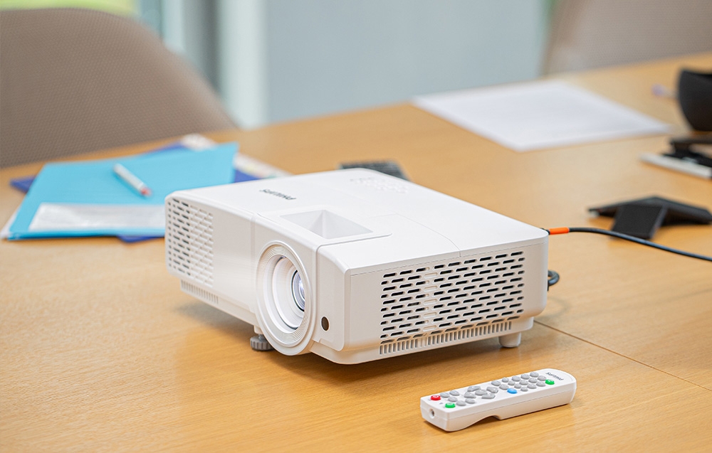 Ένας λευκός προβολέας PHILIPS βρίσκεται πάνω σε ένα ξύλινο τραπέζι δίπλα σε ένα τηλεχειριστήριο και μπλε φακέλους