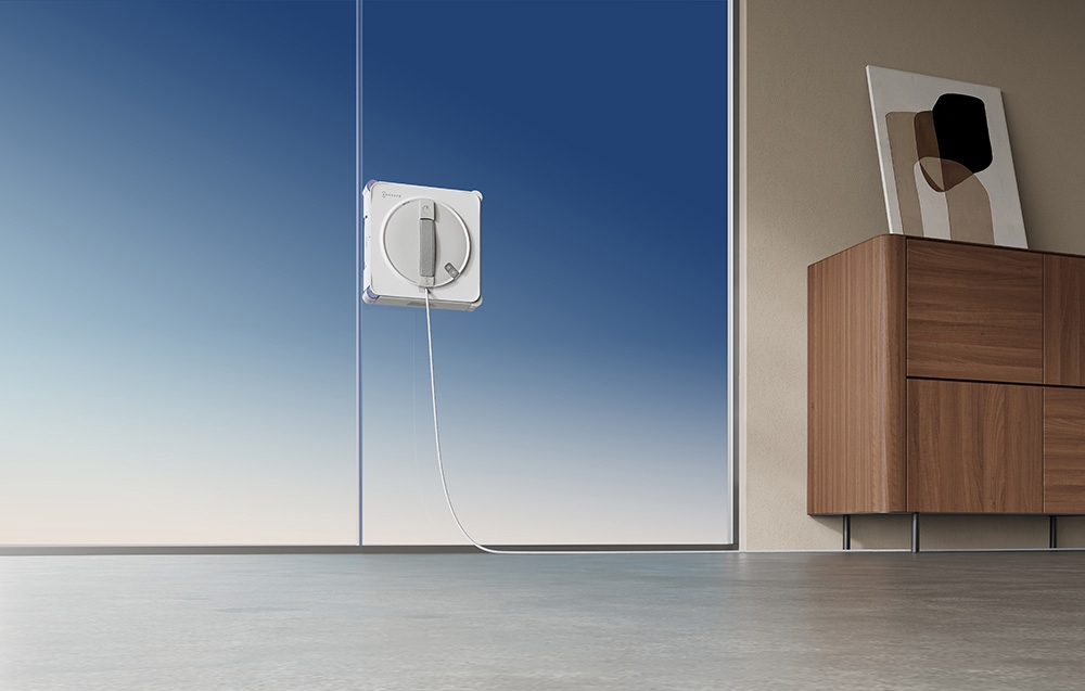 Ρομπότ ECOVACS που καθαρίζει ένα παράθυρο σε ένα φωτεινό δωμάτιο, δίπλα σε μια ξύλινη συρταριέρα με έναν πίνακα