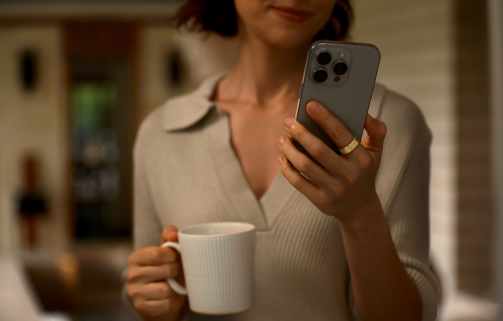 Μια γυναίκα που κρατά ένα smartphone και μια κούπα, με ένα χρυσό έξυπνο δαχτυλίδι στον δείκτη της