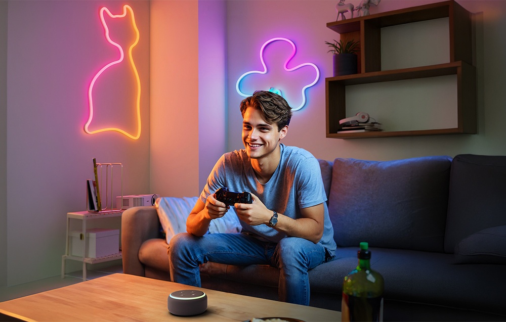 Ένας gamer κάθεται στον καναπέ με ένα χειριστήριο. Νεονικά περιγράμματα μιας γάτας και σύννεφων λάμπουν στους τοίχους.