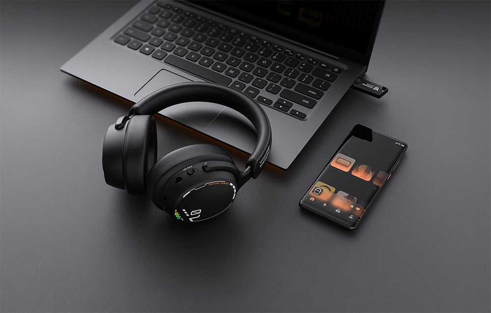 Μαύρα ακουστικά τύπου over-ear δίπλα σε ένα smartphone και έναν φορητό υπολογιστή πάνω σε ένα μαύρο τραπέζι