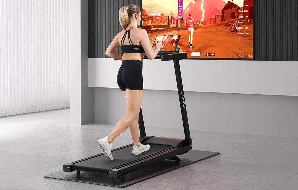 Μια γυναίκα τρέχει σε ένα μαύρο διάδρομο DeerRun μπροστά από μια μεγάλη οθόνη τηλεόρασης που προβάλλει ένα παιχνίδι τρεξίματος.
