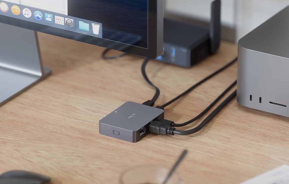 Ένας γκρι διακόπτης KVM συνδεδεμένος με καλώδια πάνω σε ένα γραφείο· κοντά του διακρίνονται μια οθόνη Mac και ένας επιτραπέζιος υπολογιστής από αλουμίνιο.