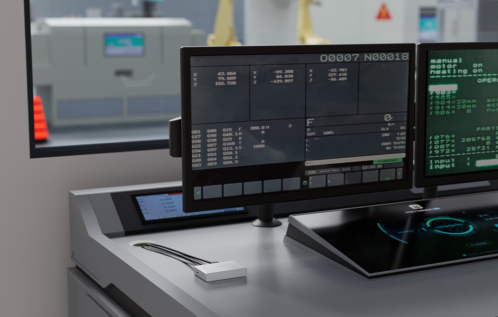 Ένας ασημένιος διακόπτης KVM πάνω σε έναν γκρι πίνακα ελέγχου σε ένα εργοστάσιο, μπροστά από οθόνες παρακολούθησης μηχανημάτων