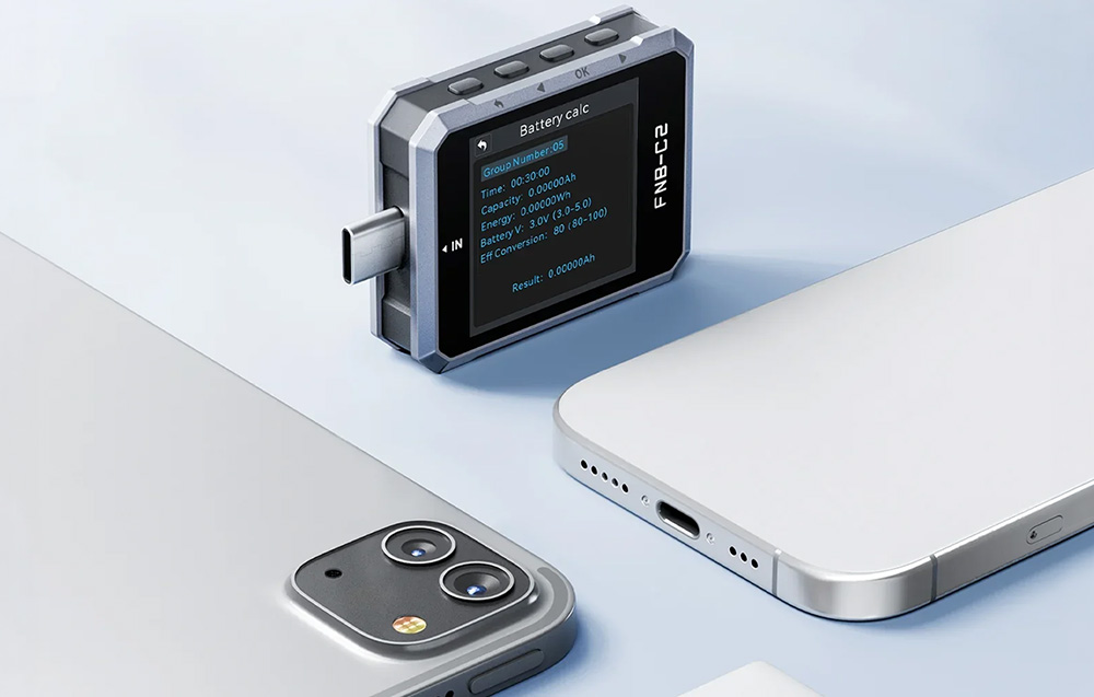 Ένας ελεγκτής USB με λειτουργία υπολογισμού μπαταρίας στην οθόνη, τοποθετημένος δίπλα σε μια θήκη smartphone πάνω σε ανοιχτόχρωμο φόντο.