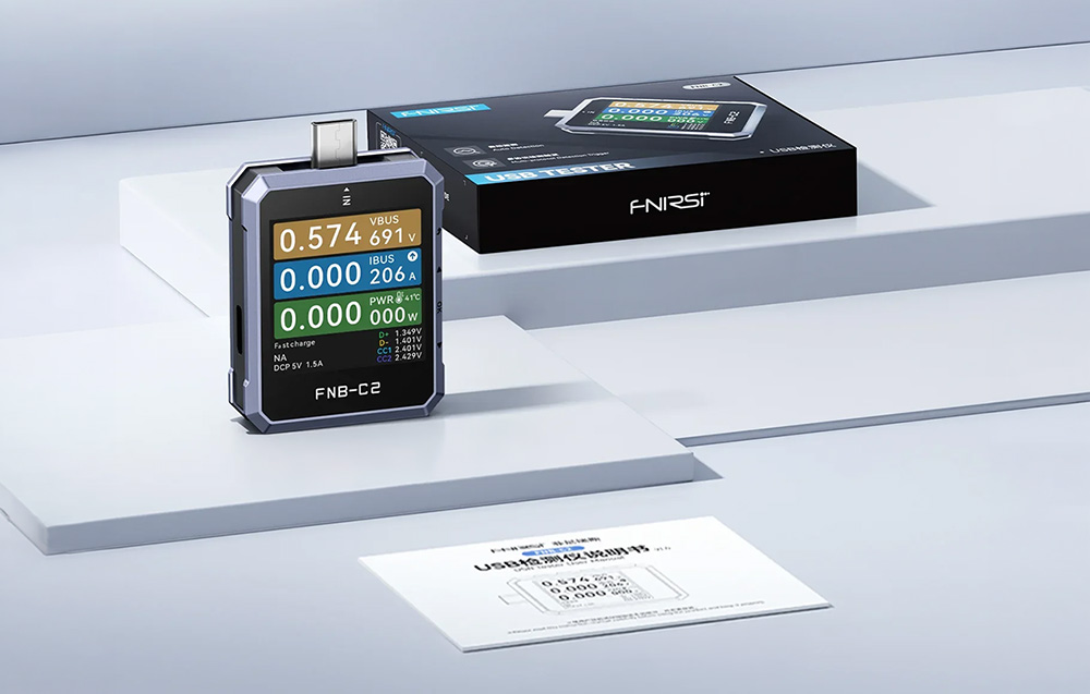 Ένας ελεγκτής USB της FNIRSI τοποθετημένος δίπλα σε ένα μαύρο κουτί και ένα έντυπο εγχειρίδιο χρήσης.