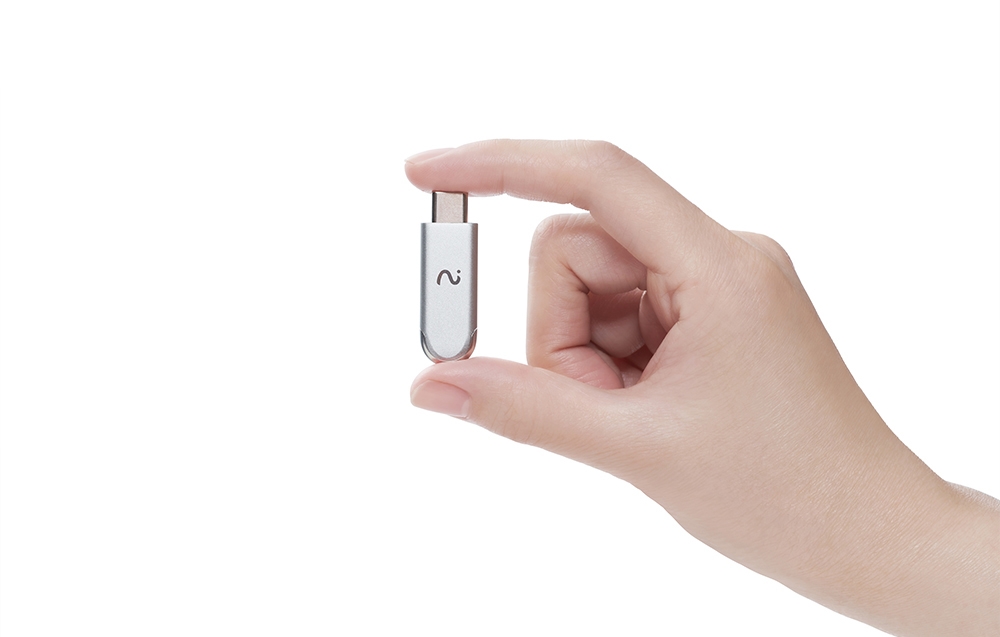 Ένα μεταλλικό dongle με στρογγυλεμένα άκρα, που φέρει ένα σύμβολο κύματος και μια μικρή οπή, κρατιέται ανάμεσα σε δύο δάχτυλα.