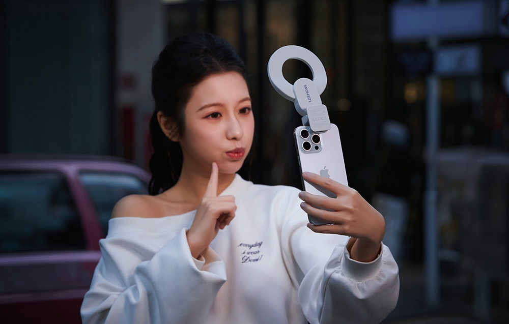 Μια γυναίκα που τραβάει μια selfie με το smartphone της, με ένα λευκό δακτυλιοειδές φως συνδεδεμένο, με φόντο έναν δρόμο της πόλης μετά το σούρουπο