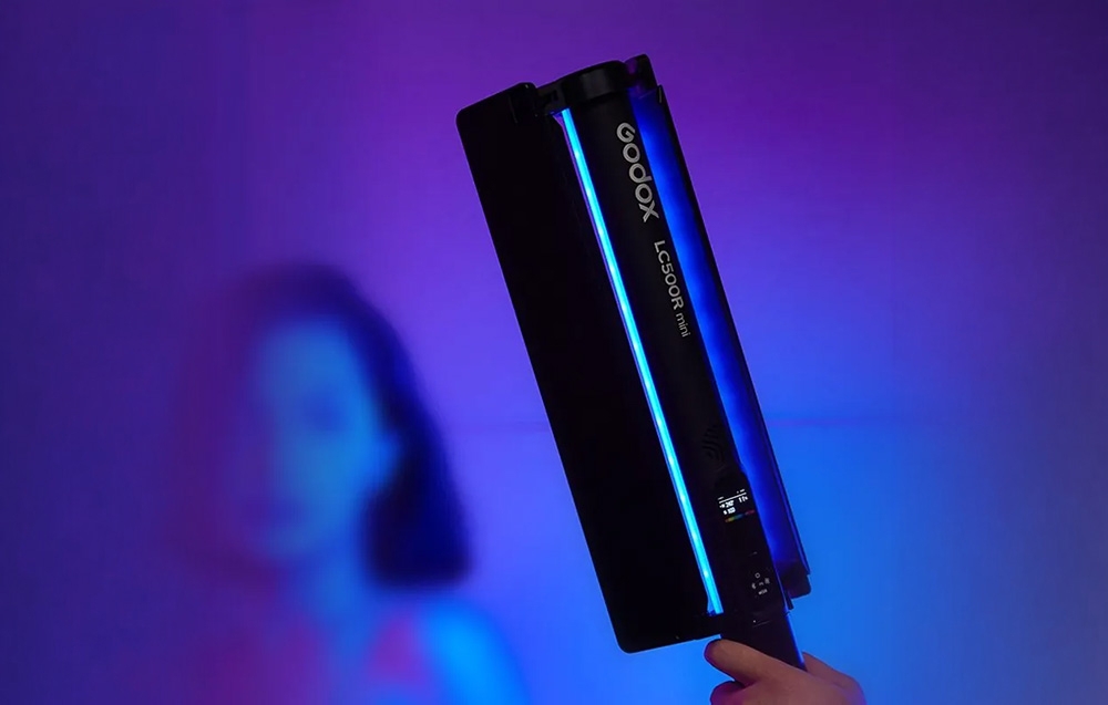 Ένα χέρι κρατά τη μπλε φωτεινή μίνι λάμπα Godox LC500R με φόντο μια θολή φιγούρα λουσμένη σε μωβ φως