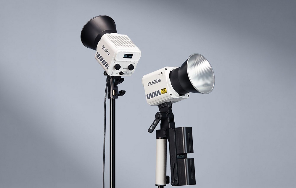 Δύο λευκά φωτιστικά Godox ML60IIBi σε τρίποδα, με ανακλαστήρες και μπαταρίες, μπροστά από φωτεινό φόντο