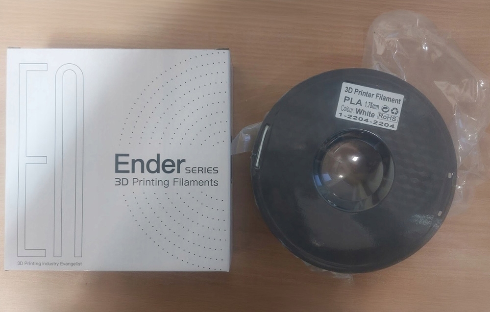 Λευκό νήμα Creality Ender-PLA σε πλαστική μεμβράνη δίπλα στο κουτί του κατασκευαστή πάνω σε ξύλινο πάγκο