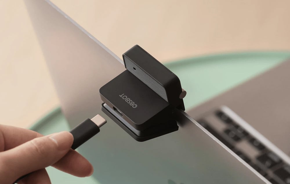 Ένα χέρι που συνδέει ένα καλώδιο USB-C σε μια μαύρη κάμερα OBSBOT τοποθετημένη στην οθόνη ενός φορητού υπολογιστή
