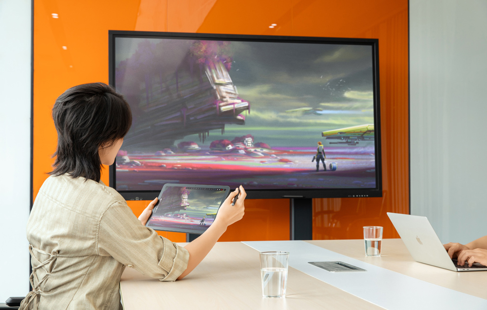 Ένα άτομο σε ένα τραπέζι κρατά ένα tablet με μια γραφίδα, και η εικόνα από την οθόνη του εμφανίζεται σε μια μεγάλη οθόνη