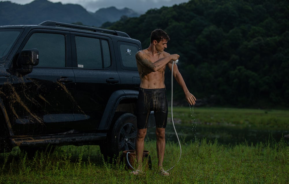 Ένας άνδρας χρησιμοποιεί το φορητό ντους Flextail Max Shower δίπλα σε ένα αυτοκίνητο στην ύπαιθρο.